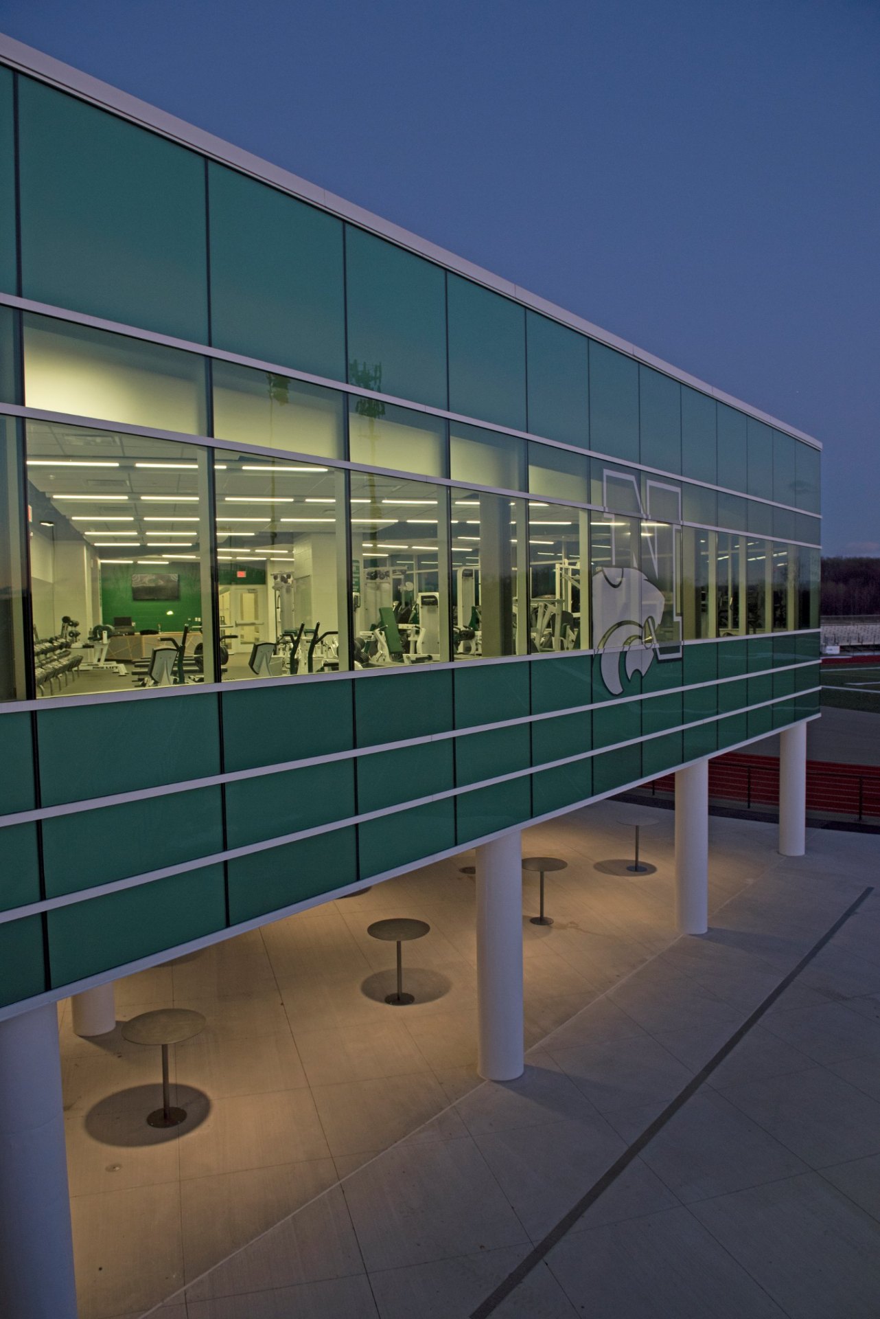 Side view of the Novi High School fitness center at dusk, Bone White Tubelite 400 curtain wall with curtain wall anchors to steel and the Wildcat logo on the glass